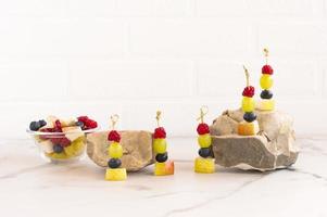 Kanapees aus saftigen Früchten am Spieß, eine Mischung aus geschnittenen Früchten in einer Glasschüssel. Zusammensetzung auf Steinen und Marmortisch. Vorderansicht. foto