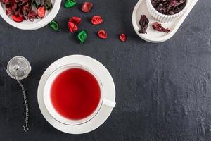 eine weiße Tasse Hibiskus-Tee auf schwarzem Hintergrund mit Rosenblättern und trockenen Hibiskus-Teeblättern. Ansicht von oben. natürliches Antibiotikum. foto