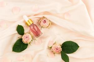 Kosmetisches Rosenöl oder ätherisches Öl in einer Flaschentropfflasche auf rosafarbenem Textilhintergrund mit Rosenknospen und Blättern. das Konzept der Naturkosmetik. foto