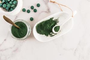 ein beliebtes Superfood für Sportler und Vegetarier. Grünes Spirulina-Pulver und Tabletten in Behältern auf weißem Marmorhintergrund. Ansicht von oben. foto