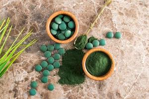 Grünes Spirulina-Pulver und Tabletten in Holzschalen auf braunem Marmorhintergrund. ein beliebtes Superfood für Vegetarier und Sportler. Ansicht von oben. foto