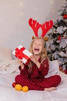 glückliches kleines blondes mädchen zu hause im pyjama für weihnachten, sortieren von geschenken und spielen mit weihnachtsspielzeug foto