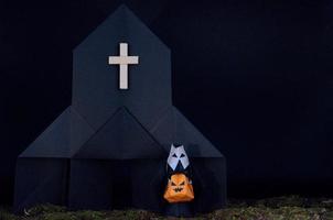 der halloween-hintergrund von origami oder papierfalten über die nonne, die kürbiskopf-jack-o-laterne vor der schwarzen kirche mit unordentlichem rasen hält foto