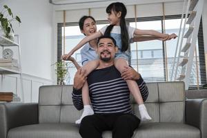 asiatische thailändische familie zusammen, vater spielt und neckt mit tochter und mutter, indem er mädchen auf den schultern im wohnzimmer trägt und hält, glückliche freizeitzeiten, schönes wochenende, wohlfühlender häuslicher lebensstil. foto