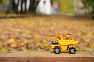 Ein kleiner gelber Spielzeuglastwagen ist mit gelben Laub beladen. Das Auto steht auf einer Holzoberfläche vor dem Hintergrund eines verschwommenen Herbstparks. Reinigung und Entfernung von Laub. saisonale Arbeiten foto