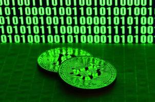 Ein Paar Bitcoins liegt auf einer Kartonoberfläche vor dem Hintergrund eines Monitors, der einen Binärcode aus hellgrünen Nullen und Einsen auf schwarzem Hintergrund darstellt. zurückhaltende Beleuchtung