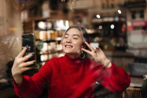 Wunderschöne Frau mit massiven Ohrringen und stilvollem hellem Pullover macht Selfie und zeigt Peace-Zeichen, en