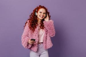 Fröhliche Frau mit roten Locken in rosafarbenem Öko-Mantel lacht, während sie über Kopfhörer Lieder hört foto