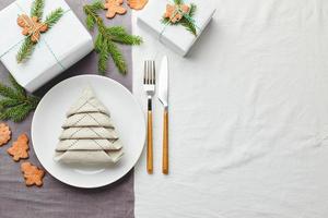 serviette in form eines weihnachtsbaums auf einem teller auf weißer tischdecke mit geschenken und dekorationen mit tannenzweigen und lebkuchenplätzchen foto