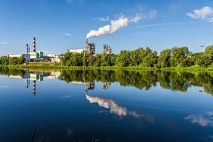 Rohre des Sägewerks des Holzbearbeitungsunternehmens mit schöner Spiegelung im blauen Wasser des Flusses. Luftverschmutzungskonzept. Industrielandschaft Umweltverschmutzung Abfall von Wärmekraftwerken foto