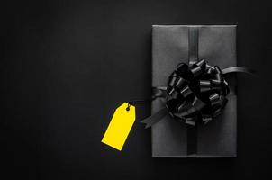 eine schwarze geschenkbox mit band und gelbem leerem preisschild setzt auf schwarzen hintergrund. schwarzer freitag und boxing day konzept.