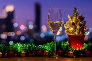 ein glas weißwein setzt auf holztisch mit weihnachtsbaum, der mit kugelverzierungen und lichtern auf buntem stadt-bokeh-lichthintergrund geschmückt ist.