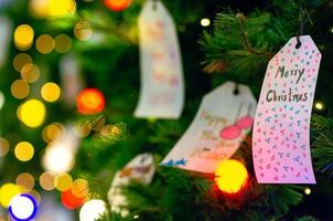 papier, das am weihnachtsbaum für feiertagsdekoration mit bunten lichtern auf hintergrund hängt. foto