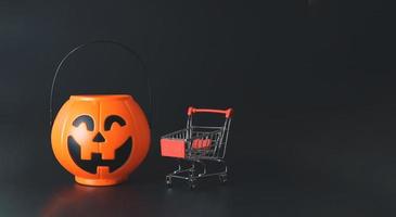 Kunststoff-Halloween-Kürbis-Eimer mit Einkaufswagen oder Trolley isoliert auf schwarzem Hintergrund mit Kopierraum. Halloween-Feiertagseinkaufskonzept. foto