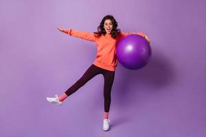 Fröhliches lockiges Athletenmädchen, das sich im lila Studio mit riesigem Fitball amüsiert. Ganzkörperaufnahme von Brunet