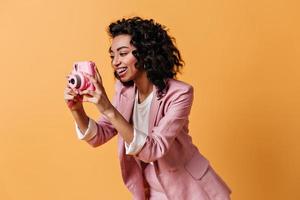 lächelnde Frau in rosa Jacke beim Fotografieren. Studioaufnahme eines trendigen Mischlingsmädchens mit Kamera.