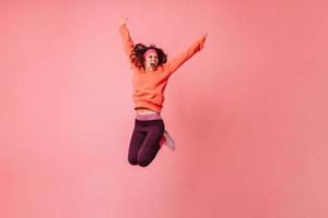 aktives mädchen in orangefarbenem kapuzenpulli und dunklen leggings, die kräftig auf rosa hintergrund springen foto