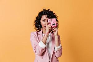 Vorderansicht einer Mischlingsfrau mit rosa Kamera. Studioaufnahme des lockigen Fotografen isoliert auf y foto