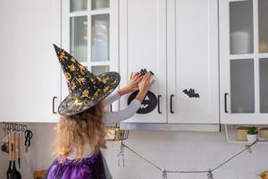 Kind schmückt die Küche zu Hause für Halloween. Mädchen in einem Hexenkostüm spielt mit dem Dekor für den Urlaub - Fledermäuse, Jack-Laterne, Kürbisse. Herbstkomfort im Haus, Küche im Scandi-Stil, Dachboden foto