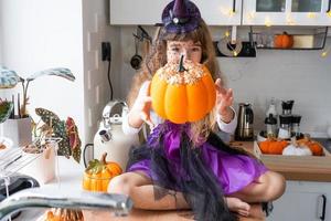 Kind schmückt die Küche zu Hause für Halloween. Mädchen in einem Hexenkostüm spielt mit dem Dekor für den Urlaub - Fledermäuse, Jack-Laterne, Kürbisse. Herbstkomfort im Haus, Küche im Scandi-Stil, Dachboden foto