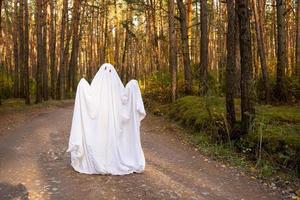 Ein Kind in Laken mit Augenausschnitt wie ein Geisterkostüm in einem Herbstwald erschreckt und erschreckt. ein nettes kleines lustiges Gespenst. Halloween Party foto