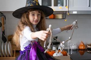 Kind schmückt die Küche zu Hause für Halloween. Mädchen in einem Hexenkostüm spielt mit dem Dekor für den Urlaub - Fledermäuse, Jack-Laterne, Kürbisse. Herbstkomfort im Haus, Küche im Scandi-Stil, Dachboden foto