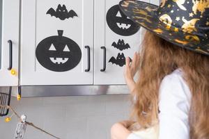 Kind schmückt die Küche zu Hause für Halloween. Mädchen in einem Hexenkostüm spielt mit dem Dekor für den Urlaub - Fledermäuse, Jack-Laterne, Kürbisse. Herbstkomfort im Haus, Küche im Scandi-Stil, Dachboden foto