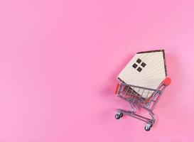 Flaches Layout des Holzhausmodells im Einkaufswagen auf rosafarbenem Hintergrund mit Kopierraum, Hauskaufkonzept. foto