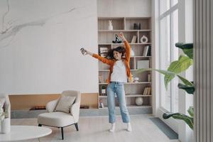 Energiegeladene Frau tanzt im Wohnzimmer mit Musik vom Smartphone im modernen Apartment-Wohninterieur foto