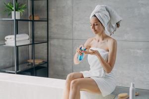 frau trägt natürliche hautpflegekosmetik auf, die im modernen badezimmer sitzt. Körperpflegebehandlung nach dem Duschen