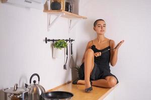 Kaukasische verführerische junge Frau im schwarzen Kleid, die auf dem Tisch in der Küche sitzt foto