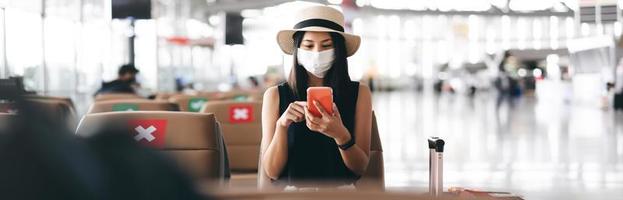 junge erwachsene touristin trägt maske für virusausbruch am flughafenterminal mit sozialem distanzierungsstuhl. foto