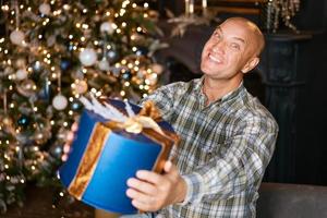 glücklicher kahlköpfiger mann, der eine schachtel mit einem geschenk auf dem hintergrund eines weihnachtsbaums hält foto