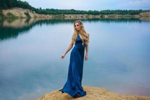 Schöne junge Frau im langen blauen Kleid steht am See foto