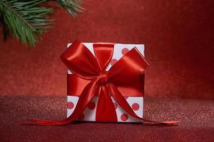 Weiße Geschenkbox mit Punkten und Schleife, Fichtenzweig auf der Rückseite vor verschwommenem funkelnden roten Hintergrund. weihnachten, neujahr. Platz kopieren foto