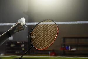 Der einzelne Badmintonspieler der Männer hält einen Schläger und einen Federball mit weißer Creme vor dem Netz, bevor er ihn auf einer anderen Seite des Platzes serviert, weicher und selektiver Fokus. foto