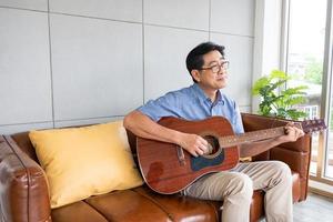 asiatischer älterer mann spielt gerne gitarre auf dem sofa innerhalb des gut innendekorationshauses. aktiver seniorenlebensstil nach der pensionierung mit glück und guter psychischer gesundheit. foto