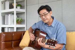 asiatischer älterer mann spielt gerne gitarre auf dem sofa innerhalb des gut innendekorationshauses. aktiver seniorenlebensstil nach der pensionierung mit glück und guter psychischer gesundheit. foto