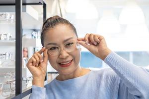 selektiver fokus auf brille. Ältere ältere asiatische Frauen lächeln und schauen in den Spiegel, während sie sich für einen schönen Brillenrahmenversuch im Optikgeschäft, Geschäft entscheiden. Sehvermögen für ältere Menschen, Gesundheitswesen. foto