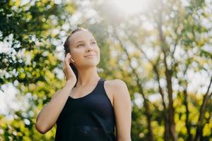 Außenaufnahme einer zufriedenen sportlichen Frau, die sich auf die Ferne konzentriert, bleibt aktiv und führt zu einem gesunden Lebensstil, der Sportkleidung trägt und Posen vor verschwommenem Naturhintergrund trägt. Sonniger Tag ist gut für das Training im Freien foto