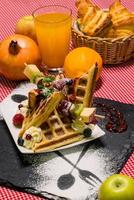Vertikale Nahaufnahme von köstlichem Dessert, Waffeln mit Obst, Croissants, Orangensaft, serviert auf schwarzem Brett foto