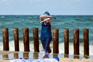 Künstlerische blauhaarige Performance-Künstlerin mit blauen Gouache-Farben, die am Strand tanzen foto