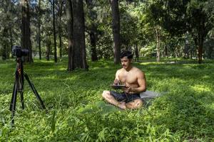 junger mann, der yoga asana praktiziert, balanciert, meditiert, während er auf einem bein auf sportmatte auf grünem gras im park steht. Verwendung des Tablets für den Online-Unterricht. foto