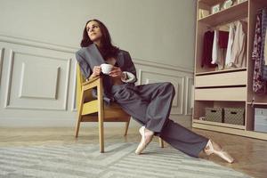 ballerina in einem grau gestreiften anzug mit einer tasse kaffee lächelnd und träumend sitzt in einem sessel foto