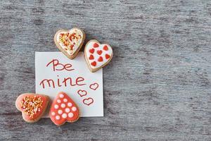 Verzierte Kekse in Herzform und Papierblatt mit der Aufschrift be mine auf dem grauen Hintergrund. Valentinstag-Konzept foto