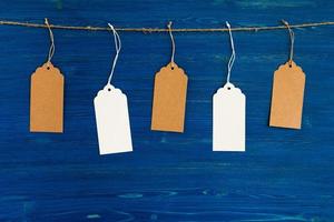 Fünf braune und weiße Preisschilder oder Etiketten aus leerem Papier, die an einem Seil auf dem blauen Holzhintergrund hängen. foto