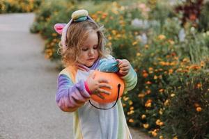 süßes kleines Mädchen in einem Regenbogen-Einhorn-Kostüm für Halloween geht, um Süßigkeiten in einem Kürbiskorb in einem Wohngebiet zu sammeln. Kind geht im Freien spazieren. Süßes oder Saures. Lebensstil. Kigurumi foto
