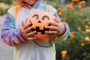 süßes kleines Mädchen in einem Regenbogen-Einhorn-Kostüm für Halloween geht, um Süßigkeiten in einem Kürbiskorb in einem Wohngebiet zu sammeln. Kind geht im Freien spazieren. Süßes oder Saures. Lebensstil. Kigurumi foto
