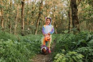 Kleines Mädchen in einem Regenbogen-Einhorn-Halloween-Kostüm und ein Dackel in einem Kleid mit einem Kürbiskorb für Süßigkeiten sitzen bei Waldsonnenuntergang auf Baumstumpf. fabelhafter wunderbarer Zauberwald. Platz für Text foto