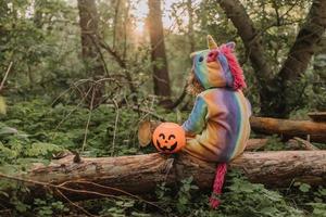 Kleines Mädchen in einem Regenbogen-Einhorn-Halloween-Kostüm mit einem Kürbiskorb für Süßigkeiten sitzt auf einem Baumstumpf bei einem Waldsonnenuntergang. ein fabelhafter wunderbarer Zauberwald. Platz für Text. hochwertiges Foto